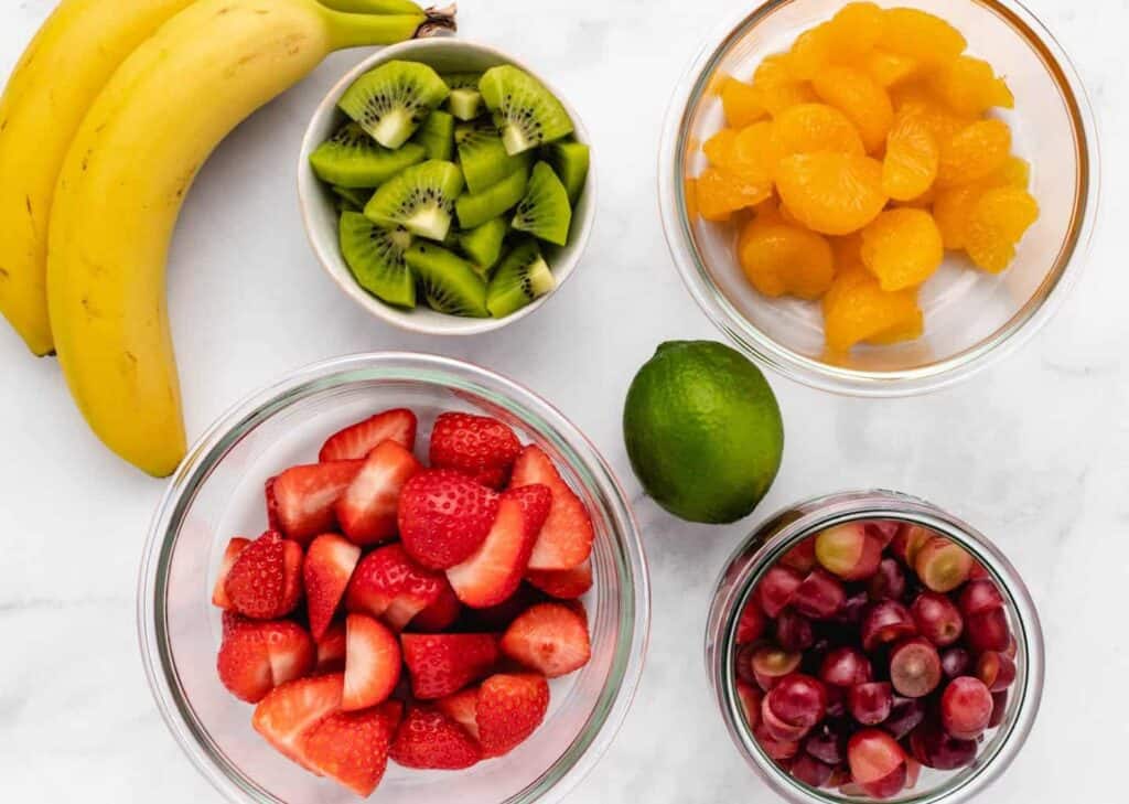 Ingredients for a fruit salad, banana, kiwi, mandarin oranges, strawberries, grapes, and lime in bowls.