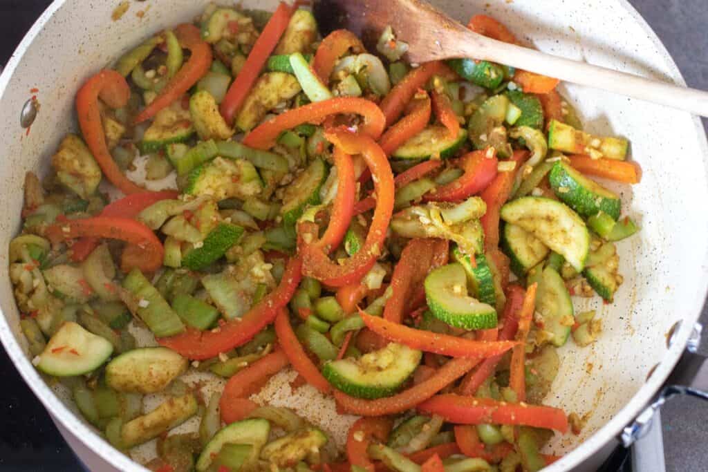 Cooked bok choy, red bell peppers, and zucchini with seasonings in a pan.