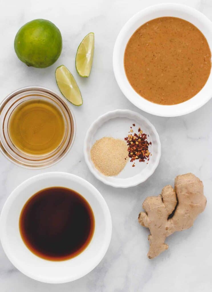 Tamari, fresh ginger, peanut butter, maple syrup, lime, and spices arranged on a countertop.