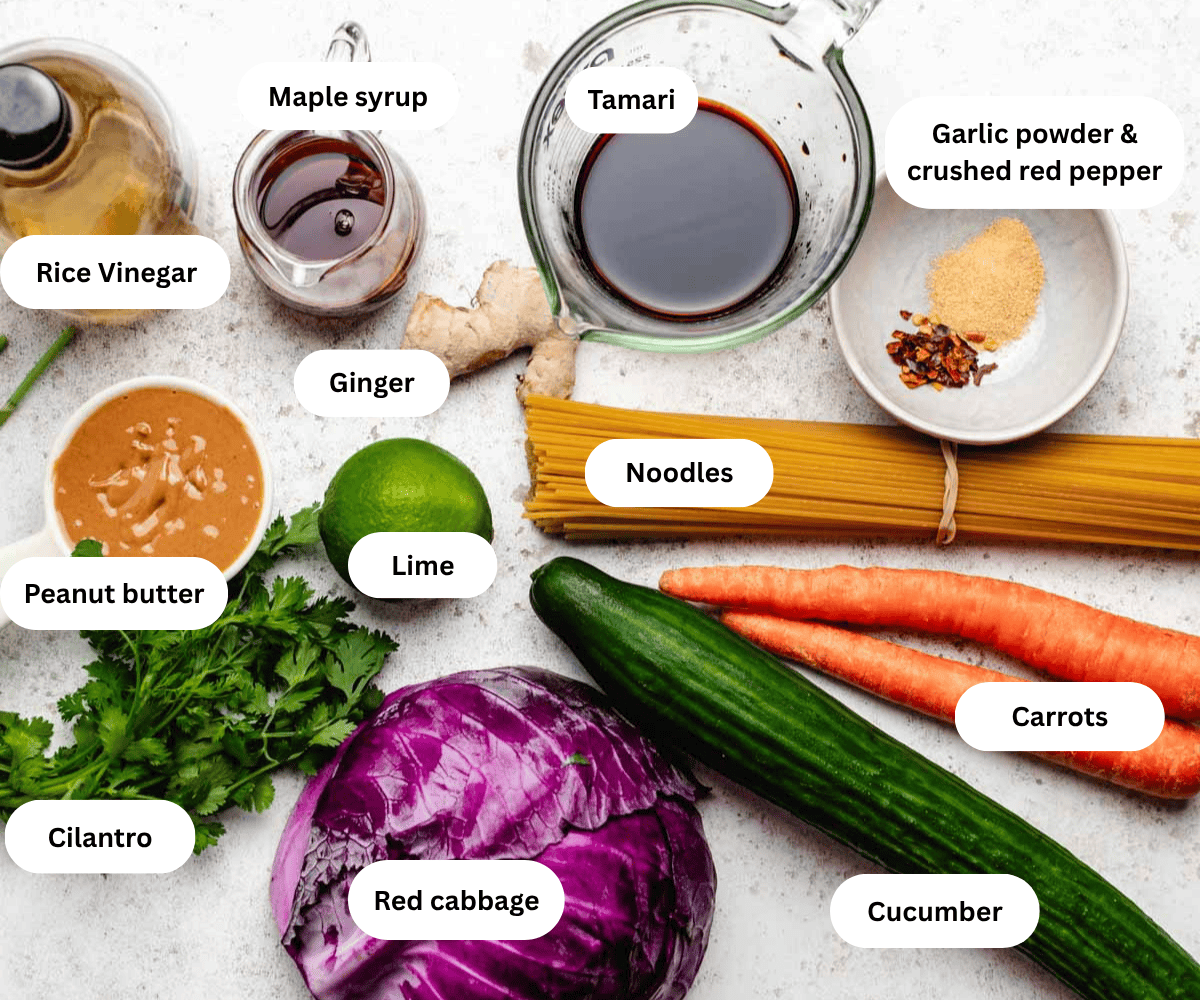 Rice vinegar, maple syrup, tamari, spices, peanut butter, lime, ginger, noodles, carrots, cucumber, cabbage, and cilantro on a table.