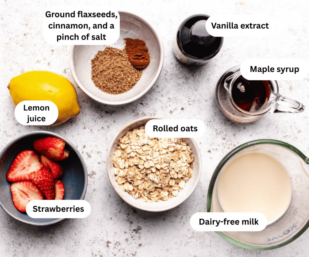 Ground flaxseeds, cinnamon, salt, vanilla extract, maple syrup, lemon, strawberries, rolled oats, and dairy-free milk on a counter.