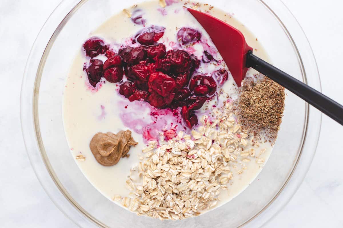 Frozen cherries, oats, flaxseeds, dairy-free milk, almond butter, and maple syrup in a bowl.