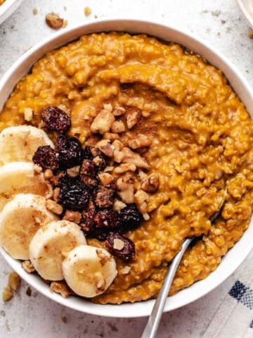 Pumpkin steel cut oats in a white bowl with sliced banana, dried cranberries, and a spoon.