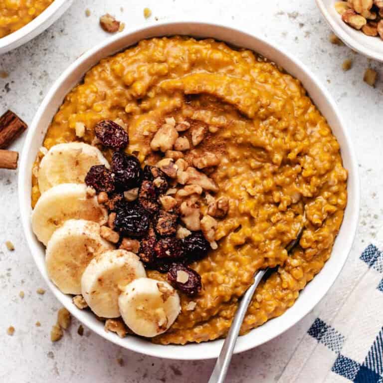 Pumpkin steel cut oats in a white bowl with sliced banana, dried cranberries, and a spoon.