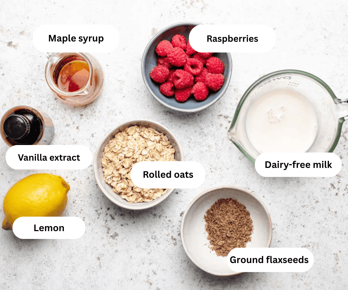 Maple syrup, raspberries, dairy-free milk, rolled oats, vanilla extract, lemon, and ground flaxseeds on a table.