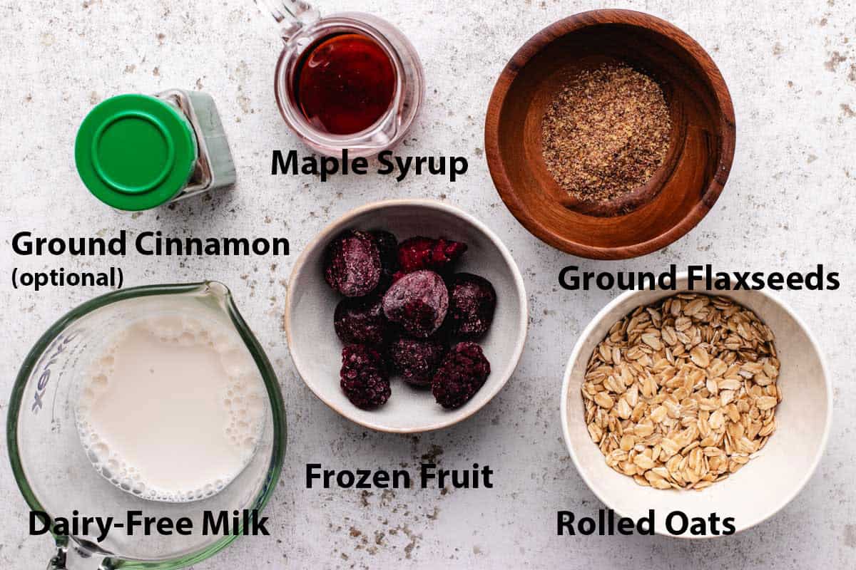 A glass of maple syrup, a bowl of ground flaxseeds, rolled oats, frozen fruit, dairy-free milk, and ground cinnamon.