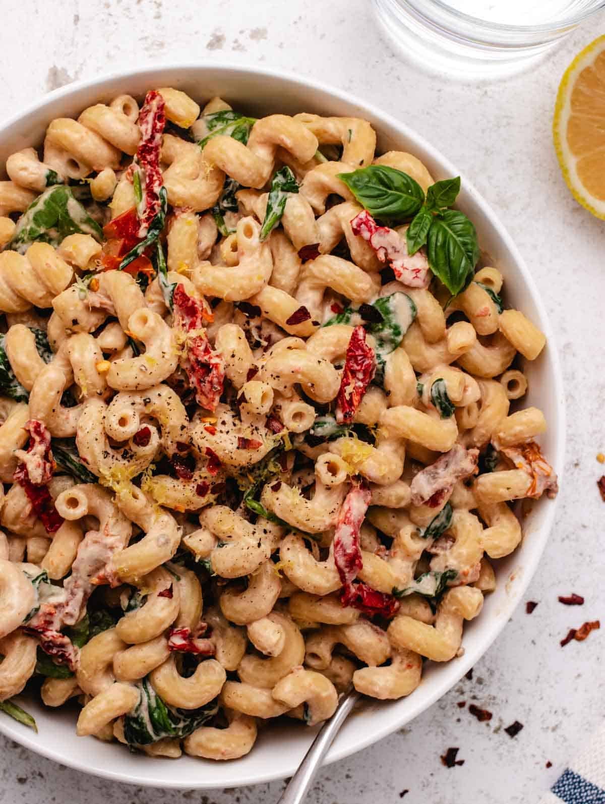 Pasta with tahini sauce in a white bowl with sun-dried tomatoes and spinach.