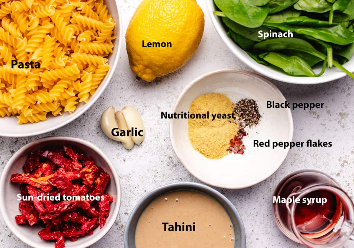 A bowl of dry pasta, lemon, fresh spinach, sun-dried tomatoes, garlic, tahini, maple syrup, nutritional yeast, red pepper flakes, and black pepper.