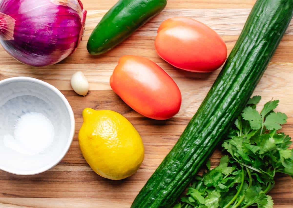 A red onion, jalapeno, lemon, tomatoes, garlic, cucumber, cilantro, and salt layed out on a counter.