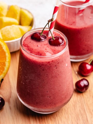 A cherry mango smoothie in a glass with a straw and two cherries.