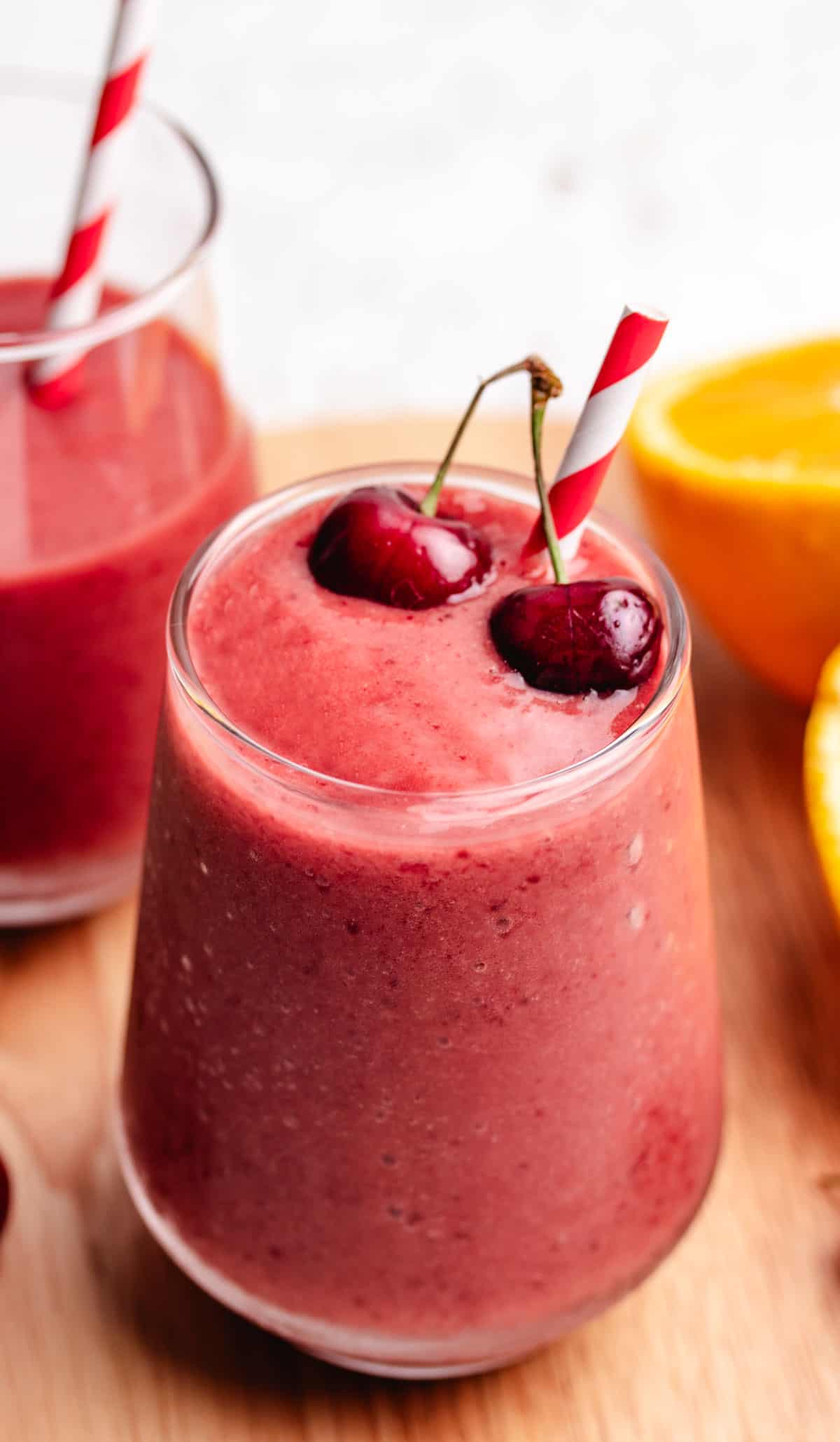 A cherry and mango smoothie in a glass with a straw and two cherries.