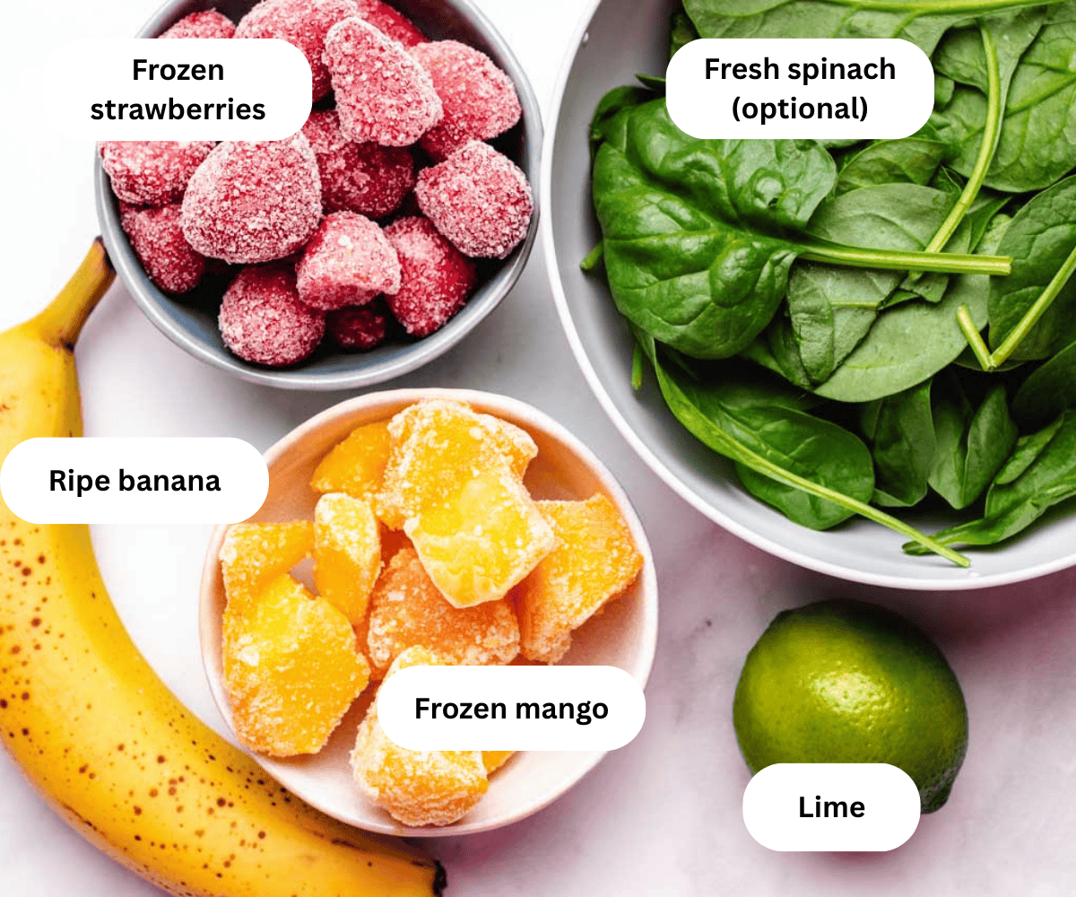 A bowl of frozen strawberries, a bowl of frozen mango, a bowl of fresh spinach, a banana, and a lime.
