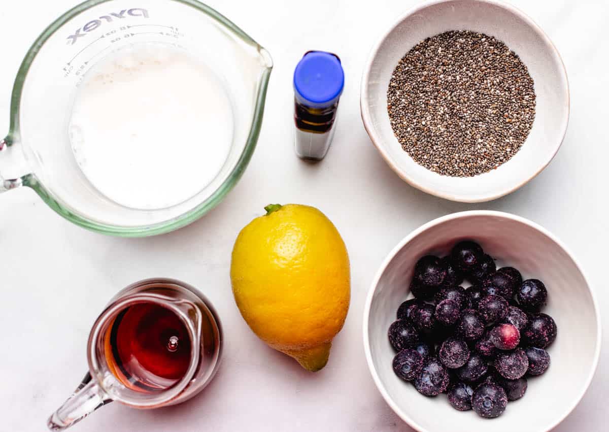 Milk in glass, whole chia seeds in a bowl, maple syrup in a jar, a lemon, and frozen blueberries in a bowl.