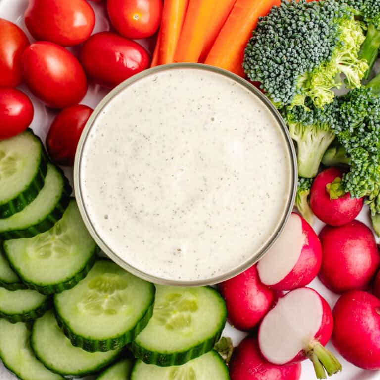 Vegan ranch dressing in a glass bowl with cucumbers, radishes, broccoli, carrots, and tomatoes.