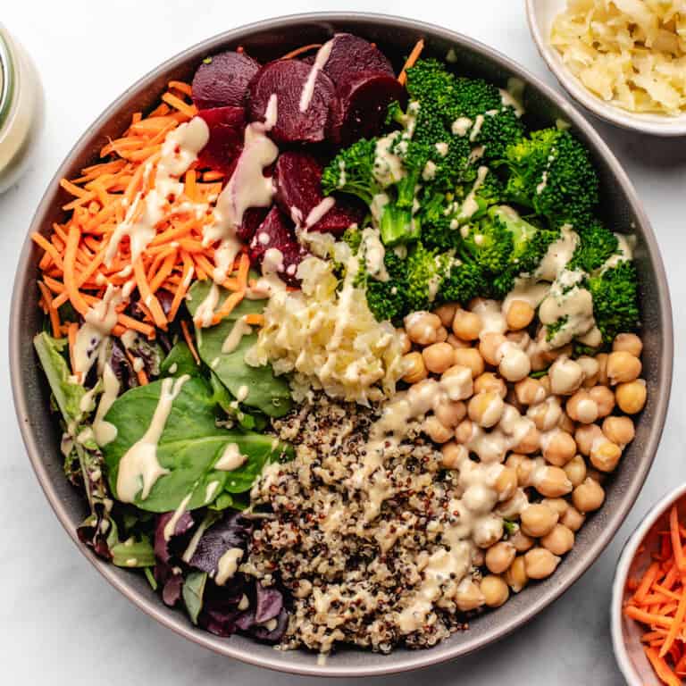 A bowl with chickpeas, quinoa, broccoli, beets, carrots, mixed salad greens, and a tahini dressing.