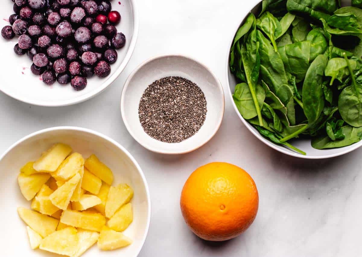 Frozen blueberries in a bowl, frozen pineapple in a bowl, chia seeds in a bowl, an orange, and a bowl of fresh spinach.