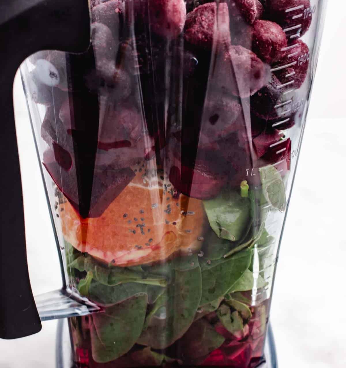 A blender with beet slices, an orange, fresh spinach, and frozen berries.