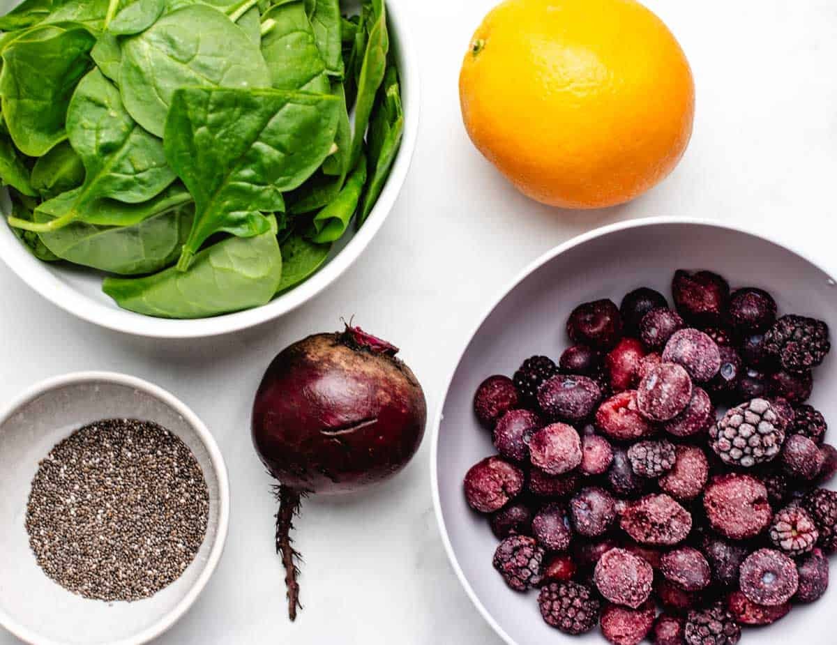 A bowl of fresh spinach, an orange, a bowl of chia seeds, a beet, and a bowl of frozen mixed berries.
