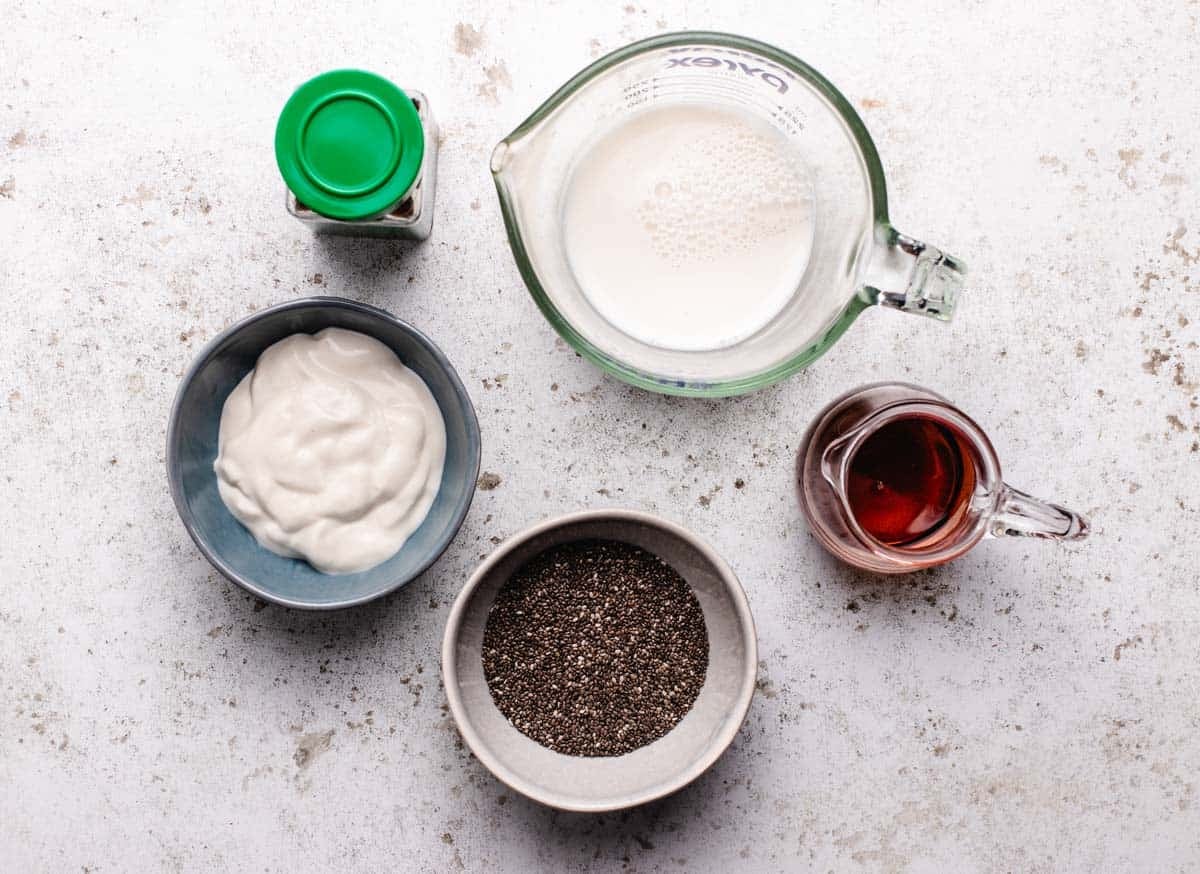 A bottle of cinnamon, a glass of dairy-free milk, a bowl of dairy-free yogurt, a bowl of chia seeds, and maple syrup.