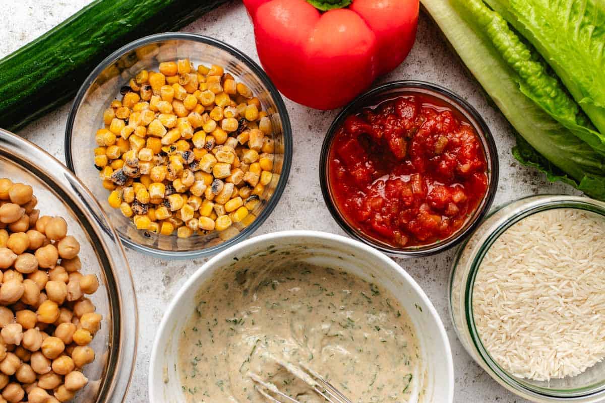 A cucumber, red bell pepper, lettuce, a bowl of corn, salsa, chickpeas, tahini lime dressing, and rice.