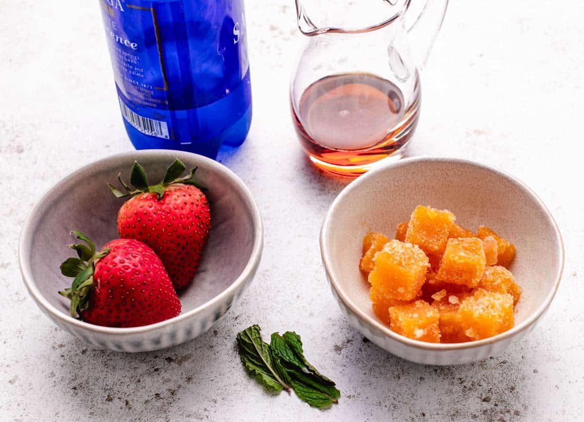 A bottle of sparkling water, maple syrup, two strawberries, mint leaves, and a bowl of frozen passion fruit puree.