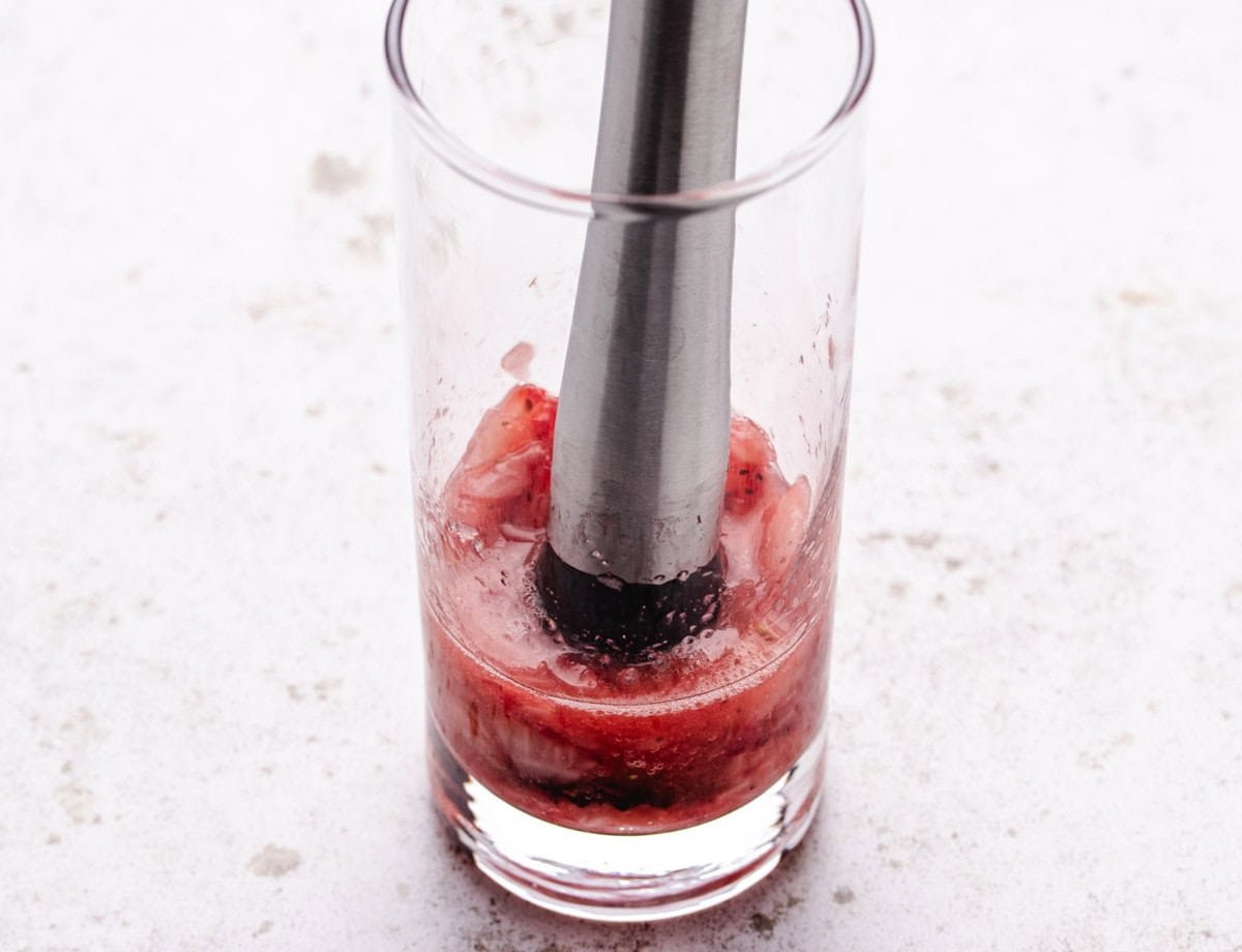 A glass of mashed strawberries with a muddler.