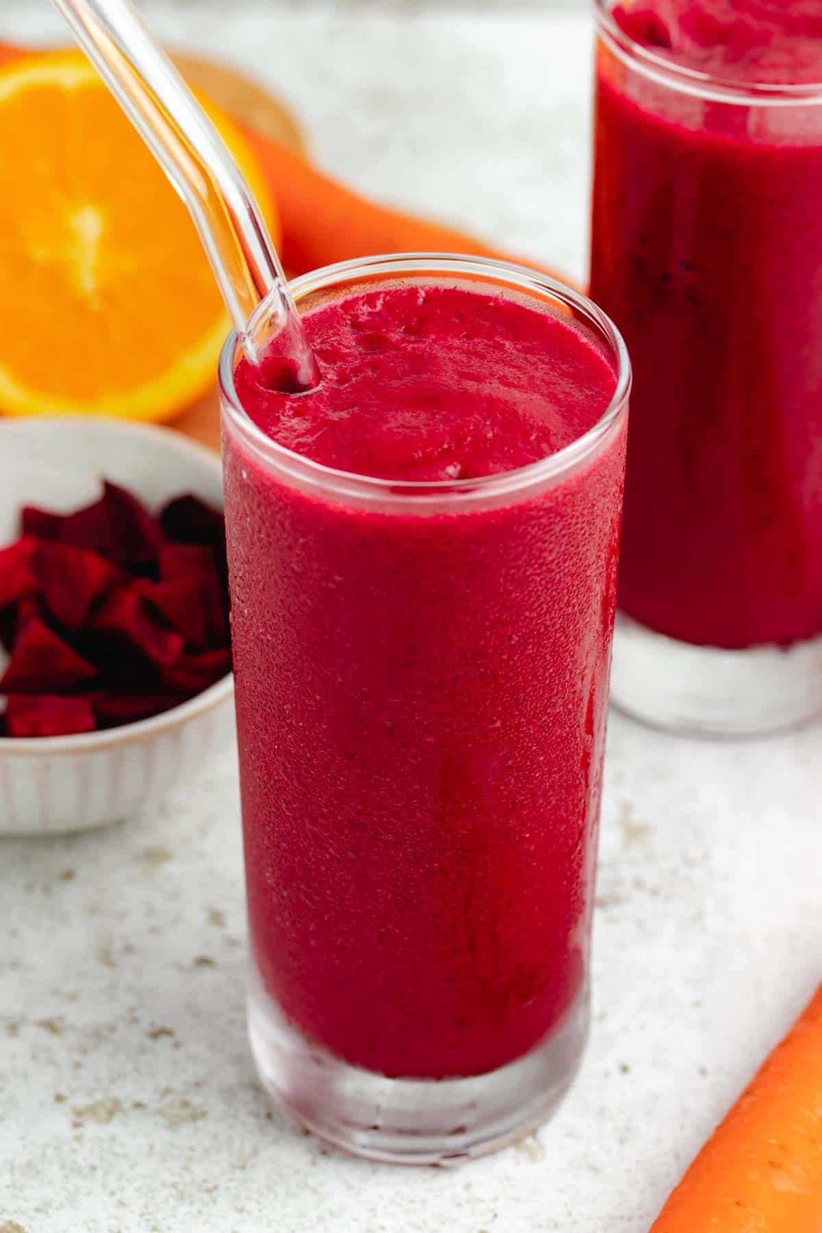 Beet and carrot smoothie in a tall glass with a glass straw and an orange and carrots in the background.