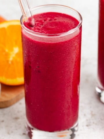 A beet carrot smoothie in a tall glass with a glass straw, with an orange and carrots in the background.