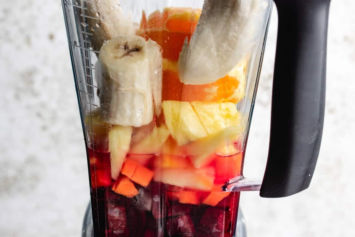 A blender with chopped beets, diced carrots, frozen pineapple, an orange, and a banana.