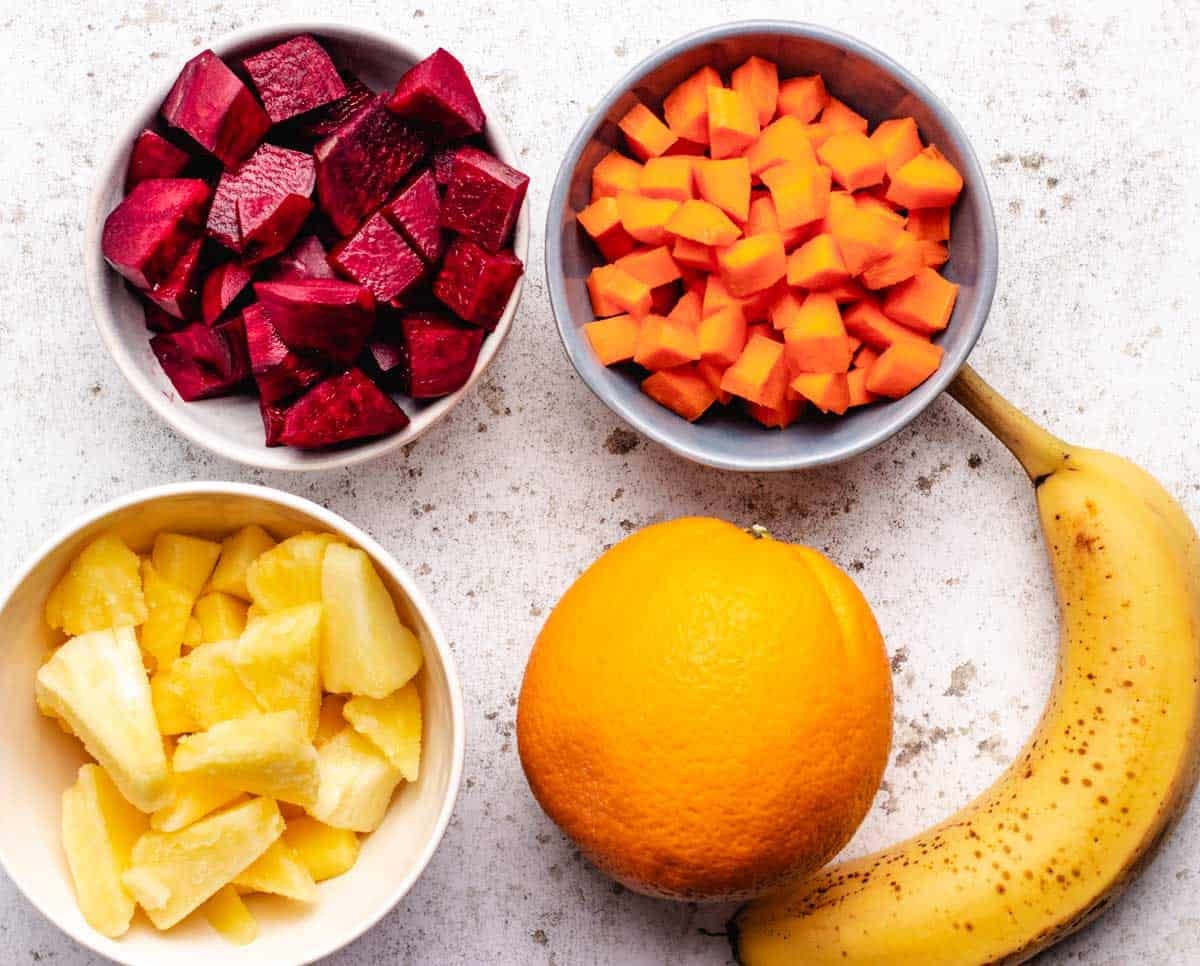 A bowl of chopped beets, diced carrots, frozen pineapple, an orange, and a banana.