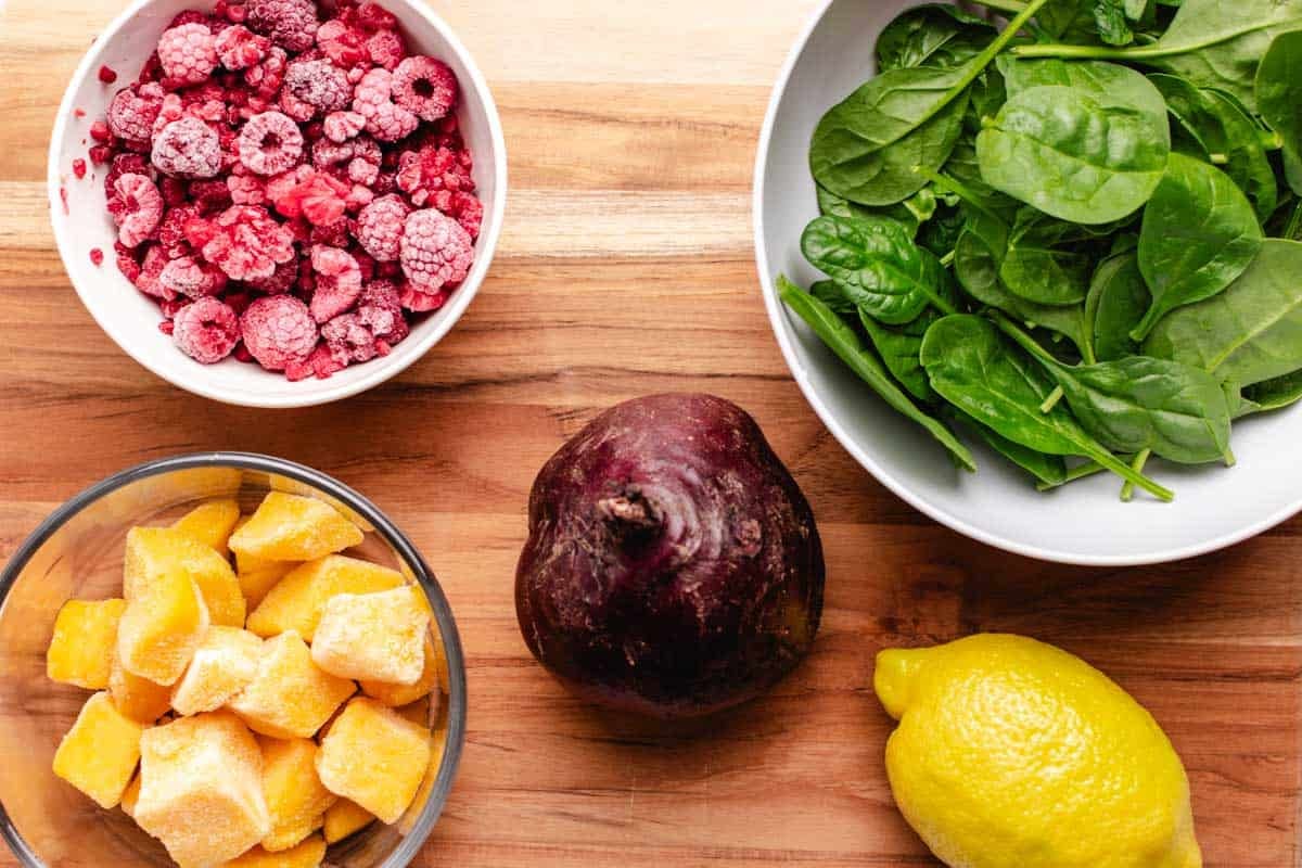 Frozen raspberries in a bowl, frozen mango, a beet, a lemon, and a bowl of spinach.