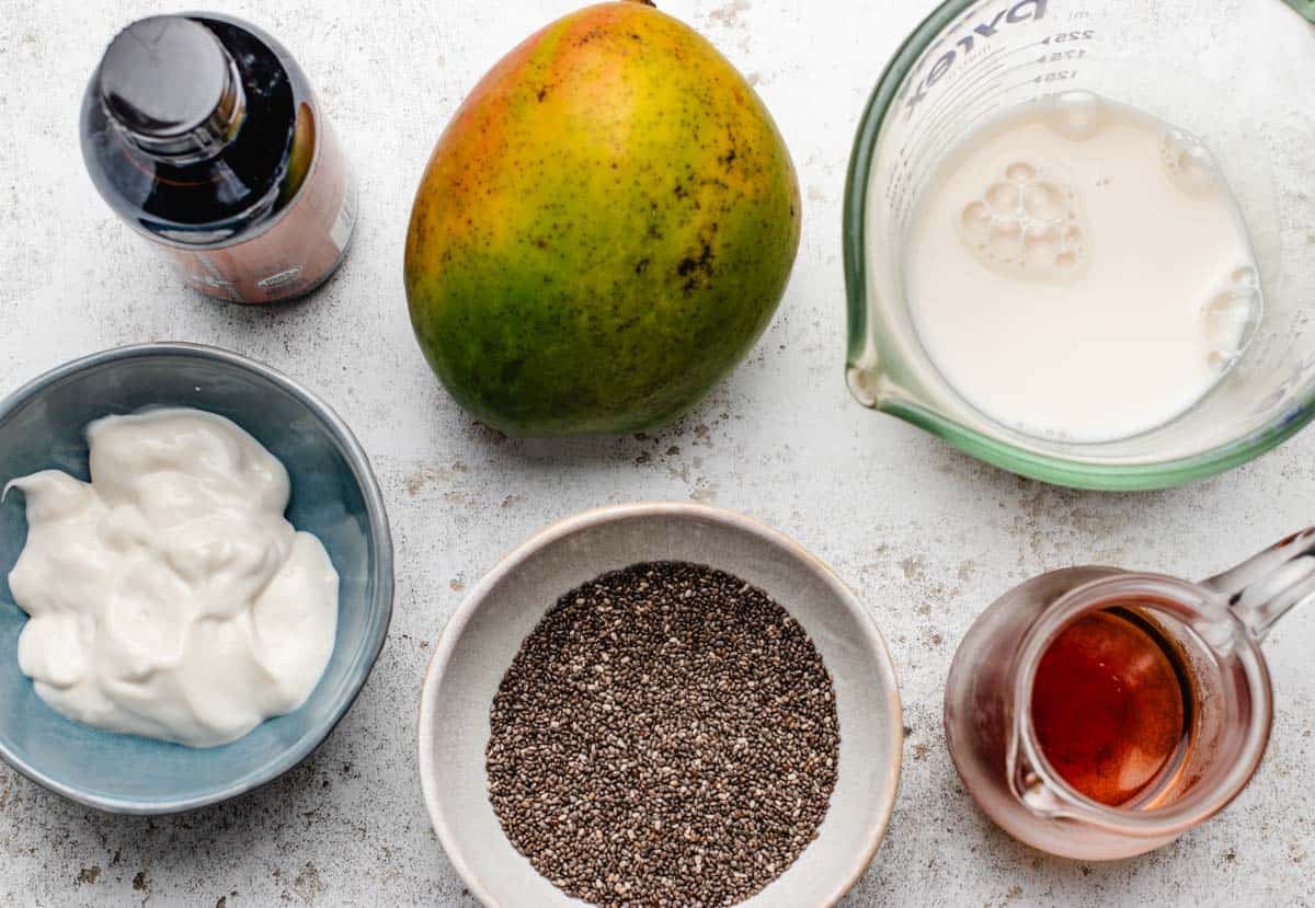 A bottle of vanilla extract, mango, dairy-free milk, a bowl of dairy-free yogurt, chia seeds, and maple syrup.