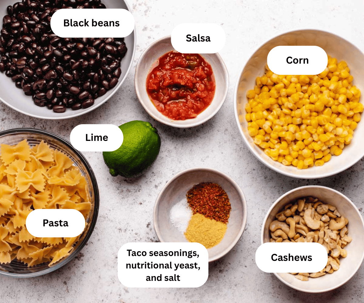 Black beans, salsa, corn, pasta, lime, seasonings, and cashews in bowls on a table.