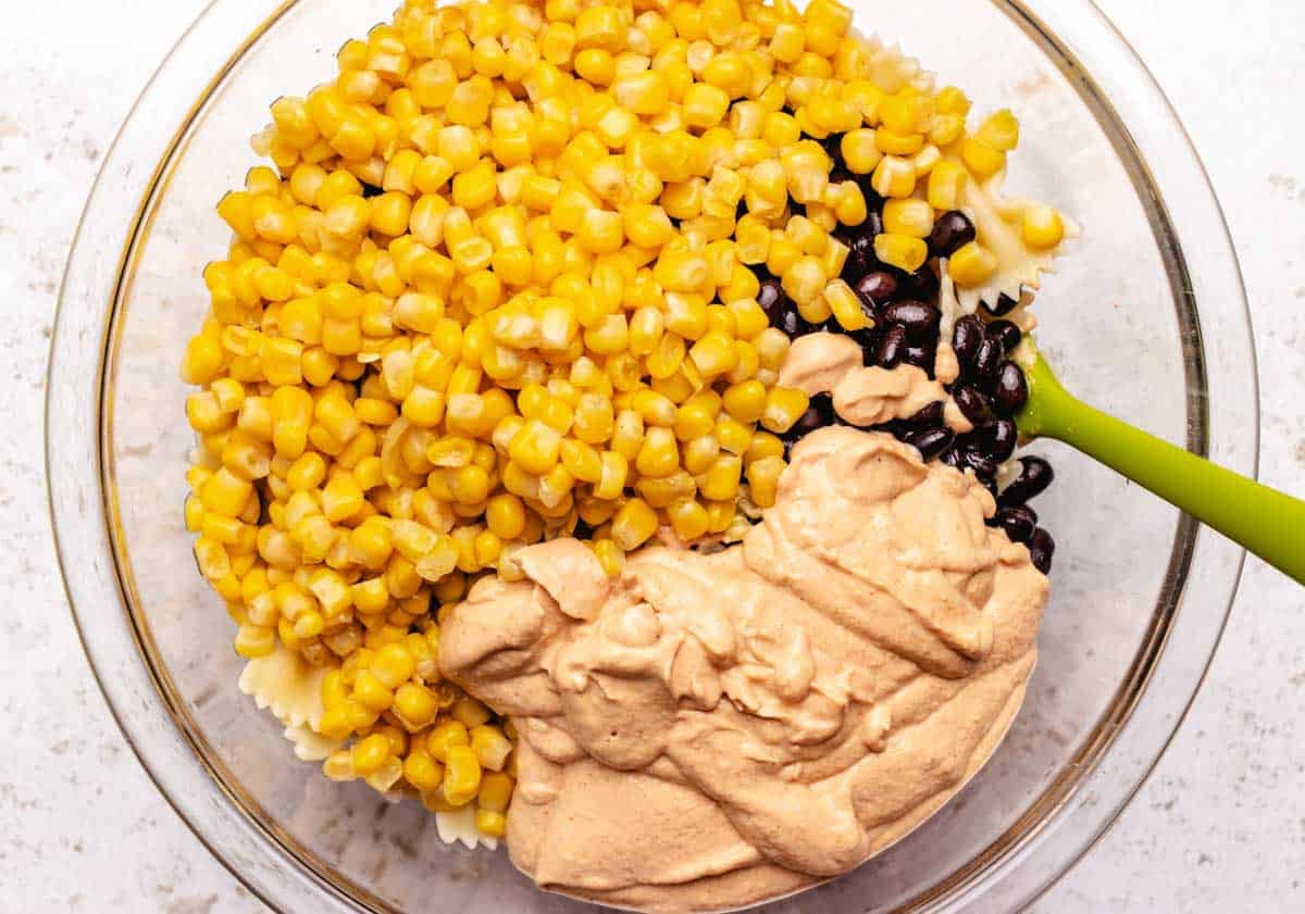 Pasta, black beans, corn, and southwest dressing in a bowl with a spatula to be tossed together.