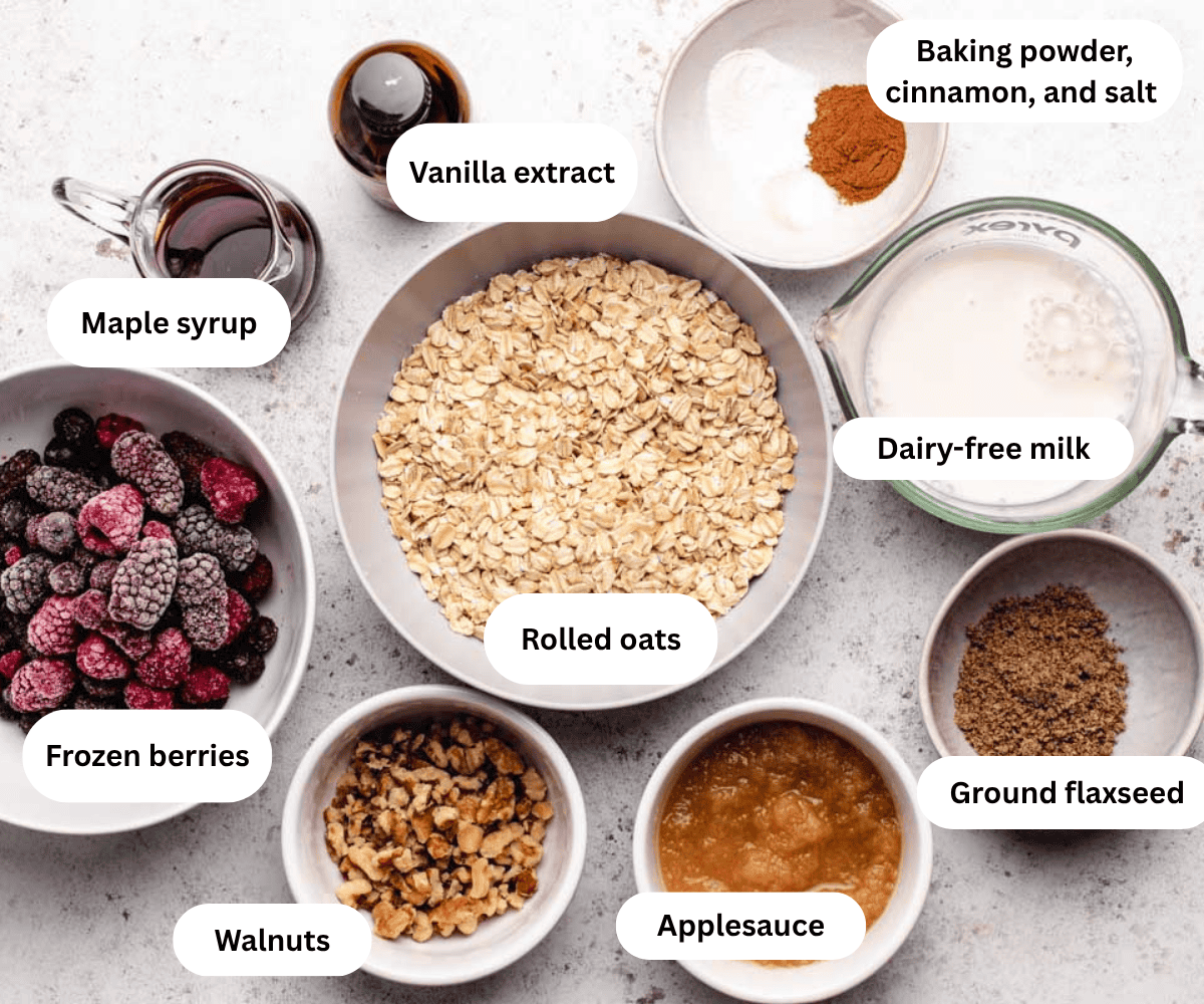 Maple syrup, vanilla extract, rolled oats, dairy-free milk ground flaxseed, applesauce, walnuts, and frozen berries in bowls.