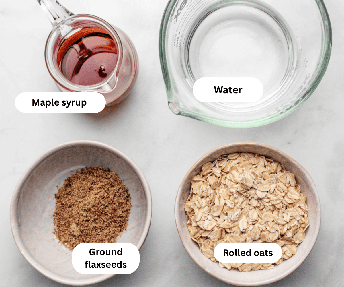 A glass of maple syrup, water, a bowl of ground flaxsees, and a bowl of rolled oats.