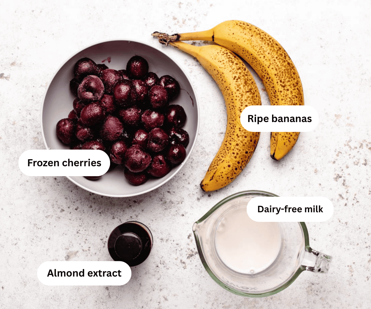 A bowl of frozen cherries, two ripe bananas, a bottle of almond extract, and a glass measuring cup of dairy-free milk.