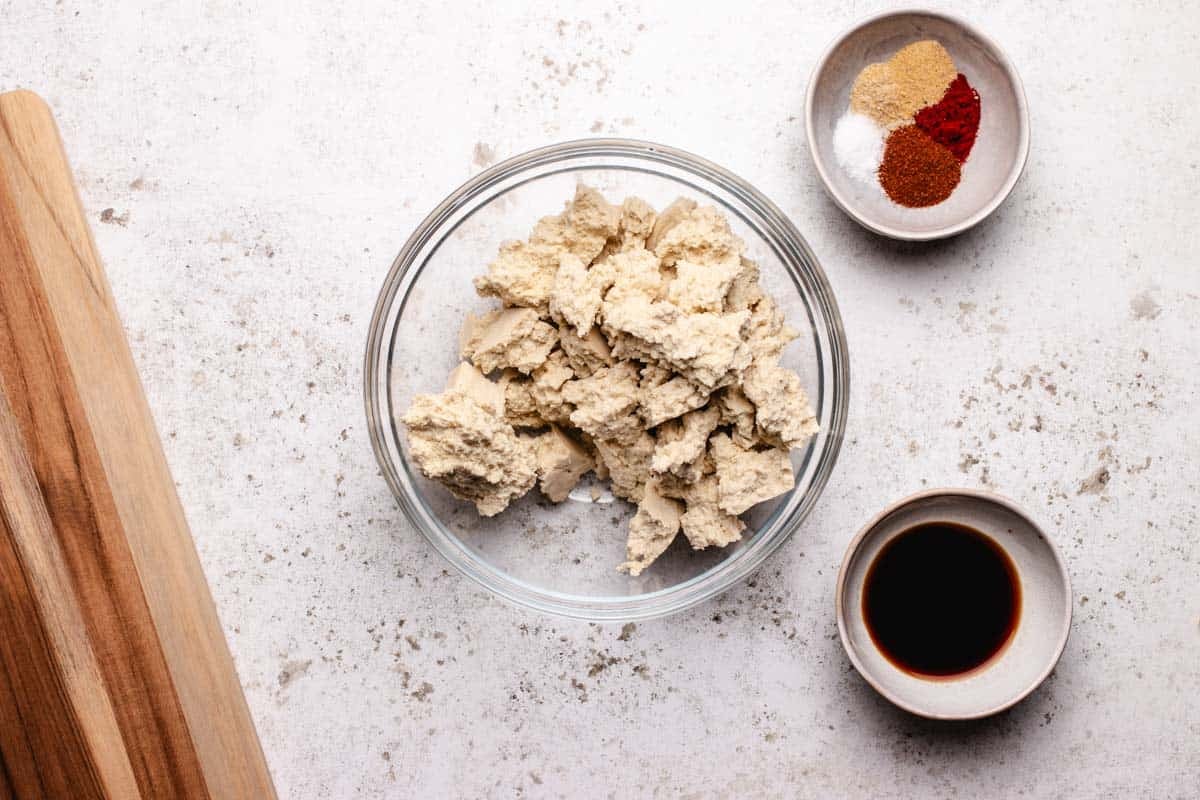 A bowl of tofu torn into pieces, a bowl of chili powder, smoked paprika, garlic powder, onion powder, and a bowl of soy sauce.