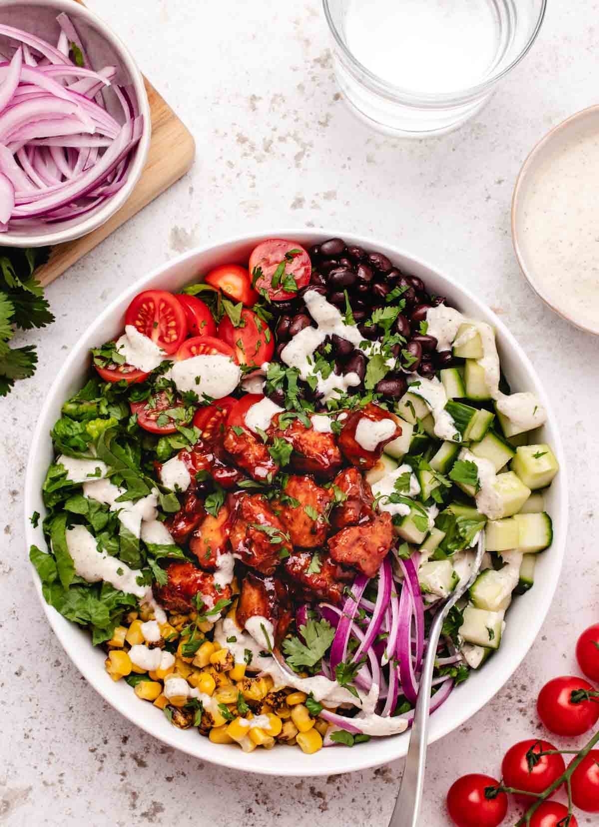 Vegan BBQ tofu salad with corn, cherry tomatoes, lettuce, red onions, cucumbers, black beans, and vegan ranch dressing in a bowl with a fork.